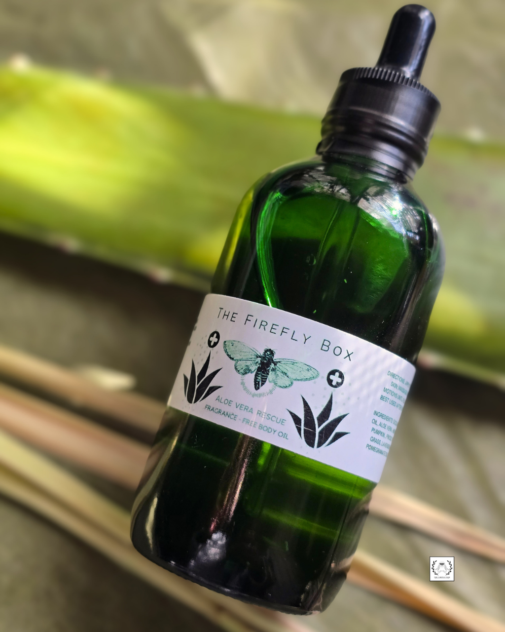 A dark green glass dropper bottle labeled 'THE FIREFLY BOX' with a black dropper cap, photographed against a blurred natural background with green leaves and tan branches.